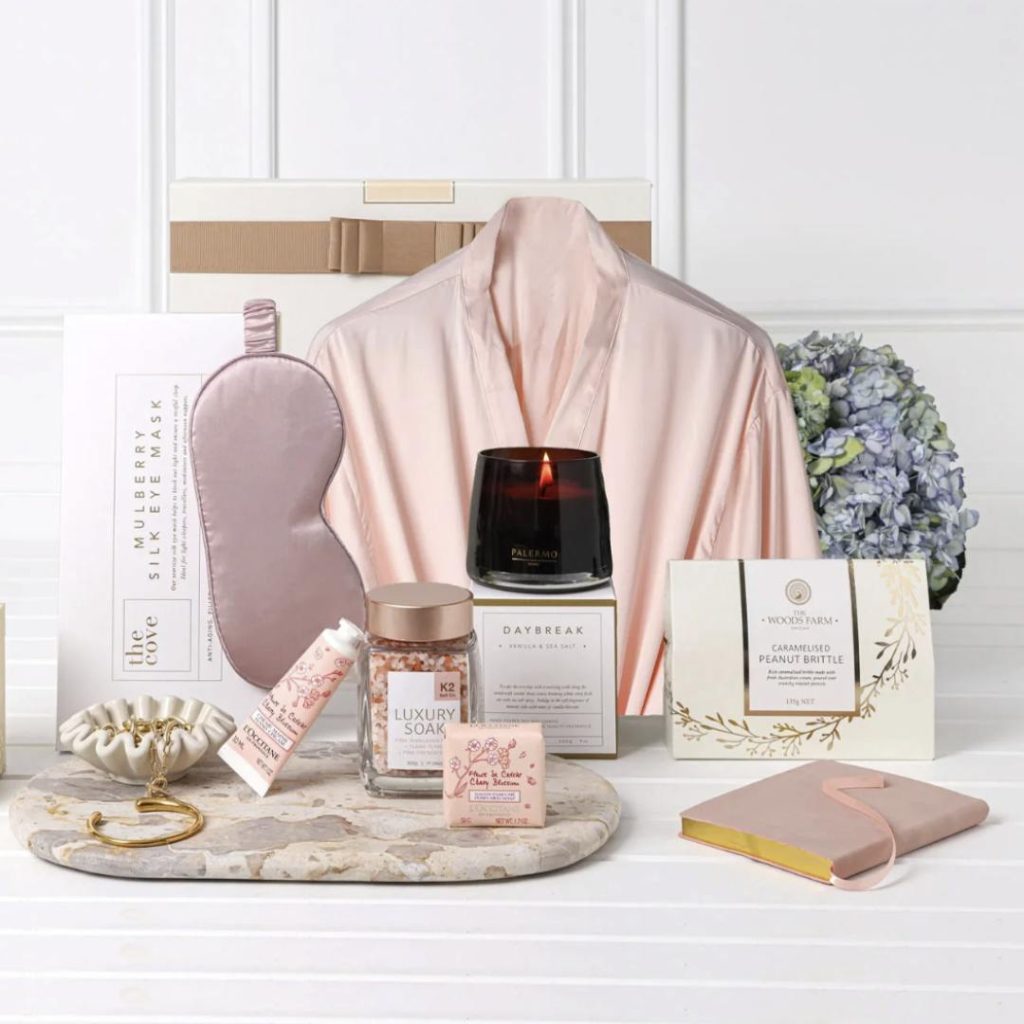 A self‑care hamper featuring a pink satin robe, sleep mask, candle, luxury bath salts, hand cream, and peanut brittle arranged on a bench