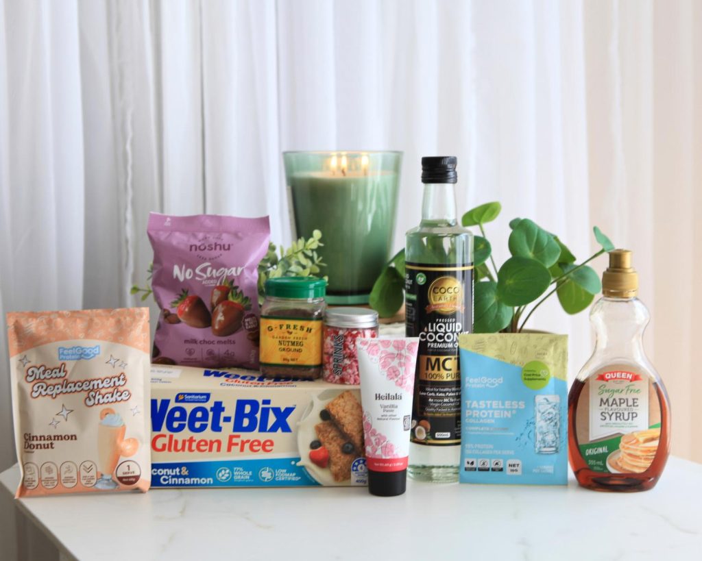 A selection of baking and protein products arranged side by side: a yellow and blue sample packet of Feel Good Protein Meal Replacement Shake; a purple packet of Noshu No Sugar Chocolate Melts; a nutmeg jar and colourful sprinkles resting on a Weet‑Bix Gluten Free box; a white tube of vanilla paste; a lit candle; a glass jar of coconut oil; a blue and yellow sample packet of Feel Good Protein Tasteless Protein Collagen; and a container of Queen Sugar Free Maple Flavoured Syrup