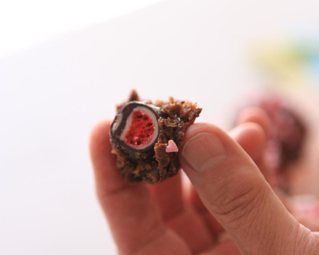 A hand holding a freeze‑dried strawberry coated in protein ball mixture, chocolate, and heart‑shaped sprinkles