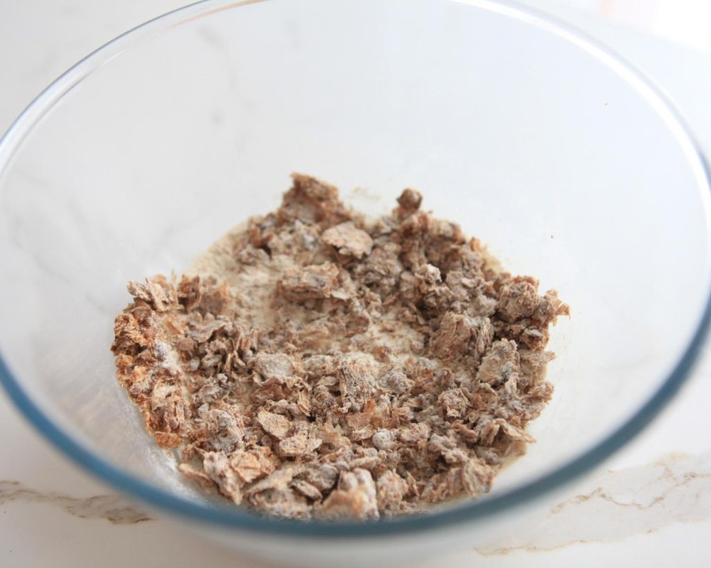 A clear glass bowl filled with a mixture of crumbled Weet‑Bix, spices, and meal‑replacement shake powder, photographed from above