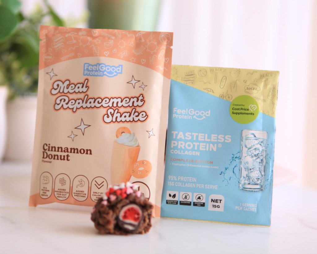 A freeze‑dried strawberry coated in protein ball mixture, chocolate, and heart‑shaped sprinkles pictured in front of a sample packet of Meal Replacement Shake by Feel Good Protein (Cinnamon Donut) and a sample packet of Tasteless Protein Collagen by Feel Good Protein