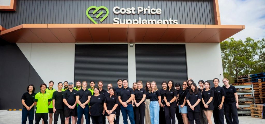 Front-facing group photo of the entire Cost Price Supplements team standing outside the new Morningside HQ warehouse, posed in front of the green and white building signage featuring a green heart and the text ‘Cost Price Supplements.’