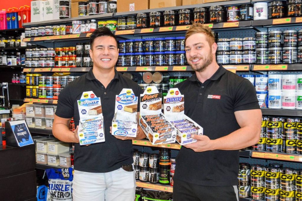 House of Supplements Founder Chris Luong and staff member Jamie posing inside the House of Supplements store in 2017, holding popular protein bar products including Bounty Protein, Milky Way Protein, Mars Protein, and Snickers Protein.