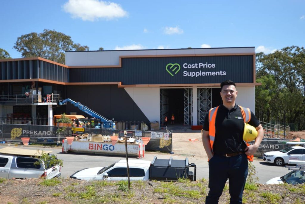 Man wearing a Cost Price Supplements polo and orange safety vest, holding a yellow hard hat while standing on a hill overlooking the 2025 construction site for the new Cost Price Supplements headquarters, surrounded by building machinery.