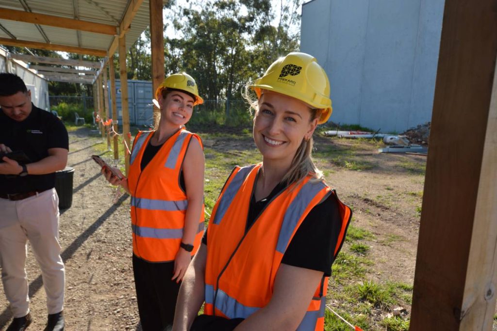 Cost Price Supplements team members Jordan and Kelsey wearing orange high‑vis vests and yellow safety helmets while touring the new Morningside headquarters in 2025.