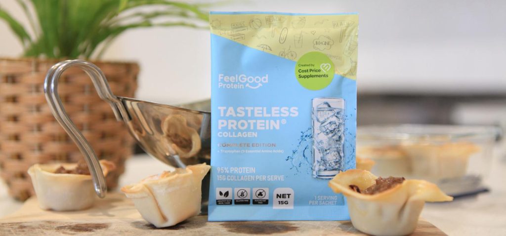 A close-up of three beef dumpling cups arranged on a wooden board, with a small silver sauce jug and a colourful Tasteless Protein Collagen sample packet beside them; a blurred plant sits in the background.