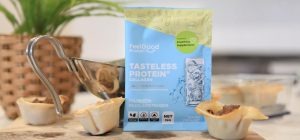 A close-up of three beef dumpling cups arranged on a wooden board, with a small silver sauce jug and a colourful Tasteless Protein Collagen sample packet beside them; a blurred plant sits in the background.