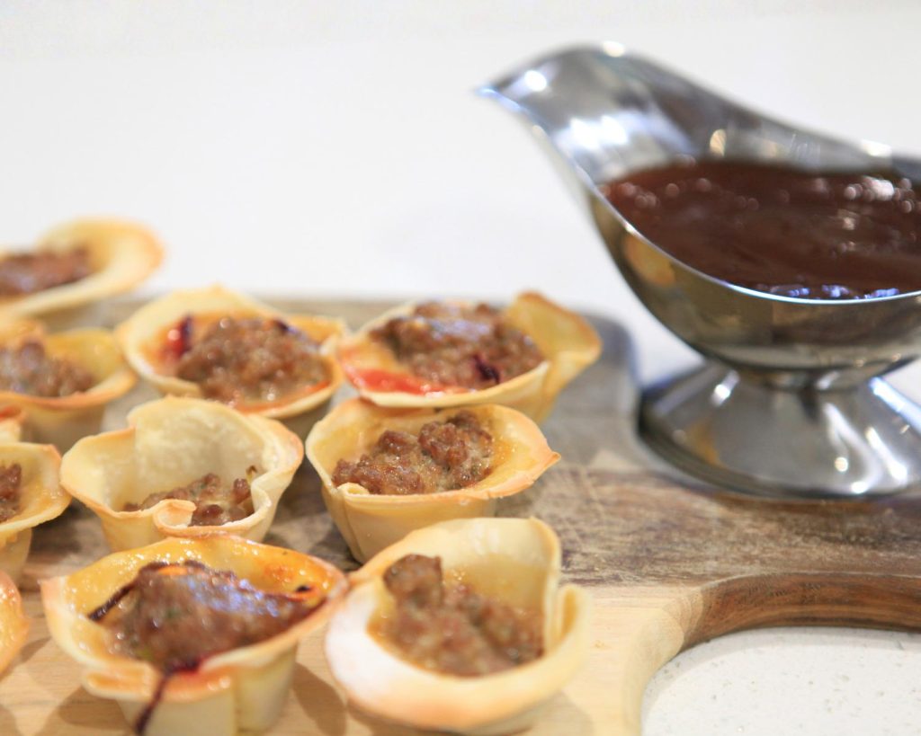 Close-up of nine Beef Dumpling Cups on a wooden board beside a small saucer of brown dipping sauce.