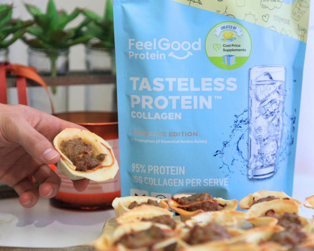 A hand lifting a Beef Dumpling Cup from a wooden board where more cups are arranged in rows, with a colourful Tasteless Protein Collagen packet in the background on the benchtop.