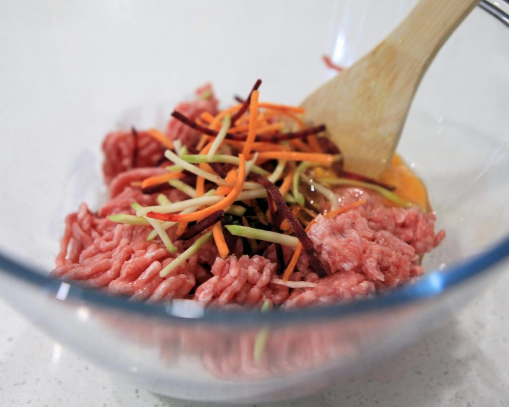 Close-up of raw beef mince in a clear glass mixing bowl, topped with a small mound of beetroot slaw, ready to be combined for the dumpling cup filling.