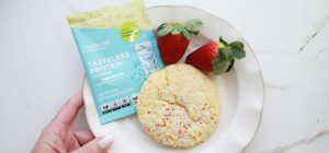Overhead view of a hand holding a white plate with a light yellow cake‑mix cookie topped with rainbow sprinkles, two red strawberries in the top left corner, and a multicoloured Feel Good Protein sachet in the top right corner.