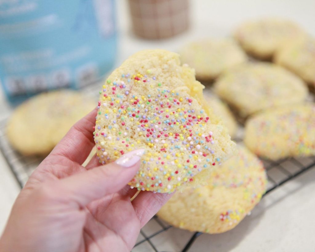 A hand holding a baked cake mix cookies coated in rainbow sprinkles in front of a blurred background. 