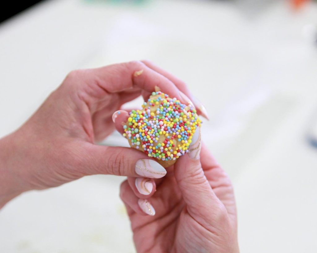 Two hands holding a half‑sphere of uncooked cookie dough coated in rainbow sprinkles against a plain white background.