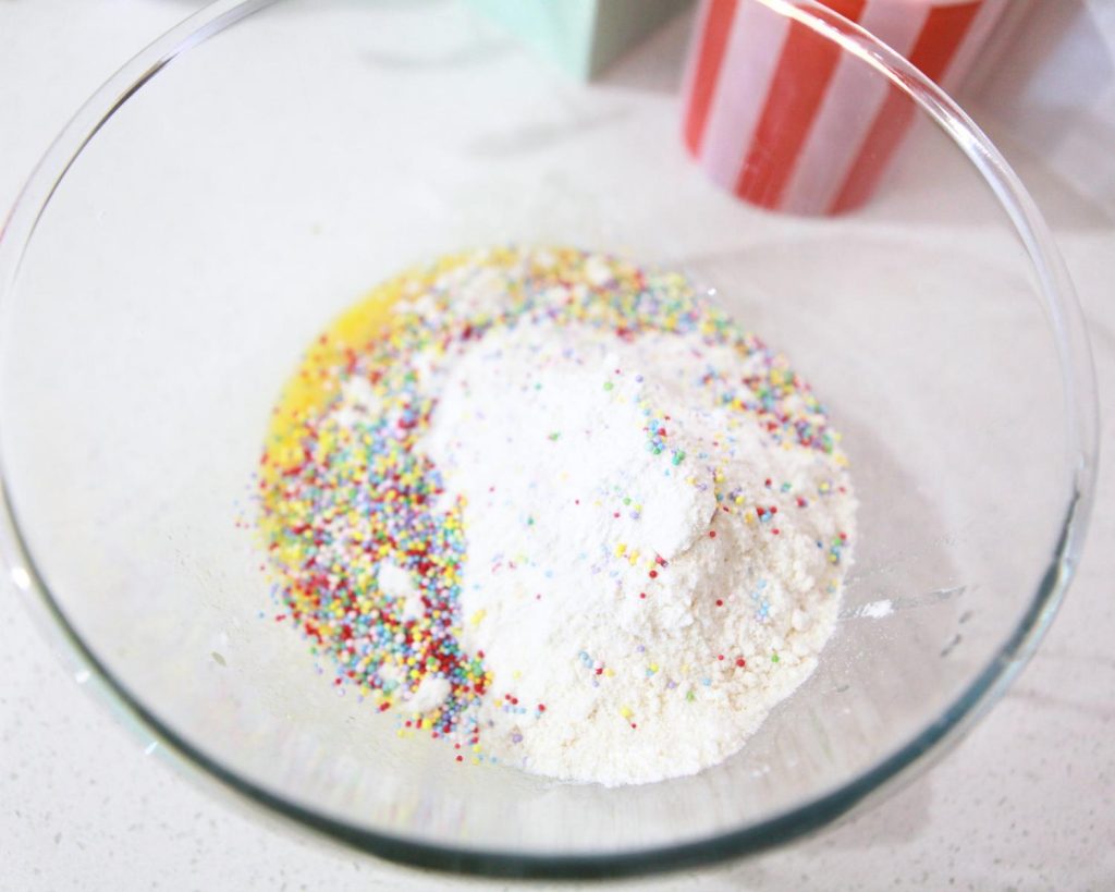 Close-up of dry cake mix cookie ingredients in a clear glass mixing bowl, topped with a small mound of 100's and 1000s sprinkles.