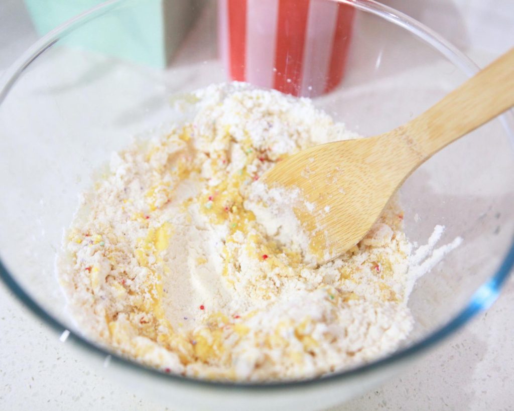 Close-up of all the mixed cookie ingredients mixed in a clear glass mixing bowl with a wooden spoon propped on the side.