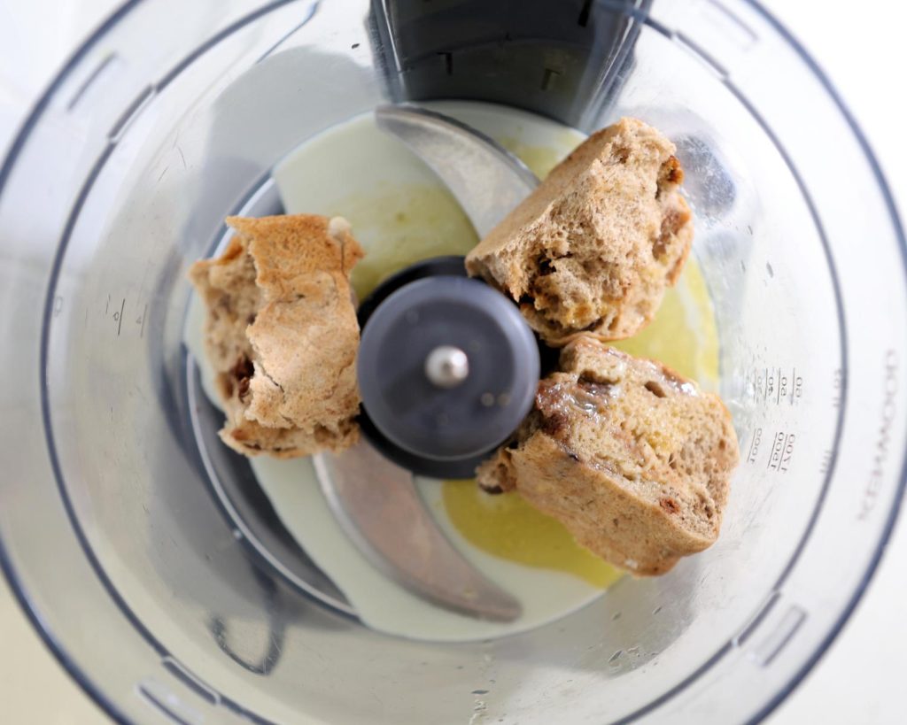 Overhead view of a food processor bowl filled with torn hot cross buns, melted butter and almond milk sitting around two silver blades.