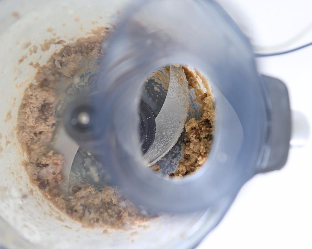 Overhead view of a food processor with a lid on, showing the bowl filled with chunky, blended ingredients sitting around two silver blades.