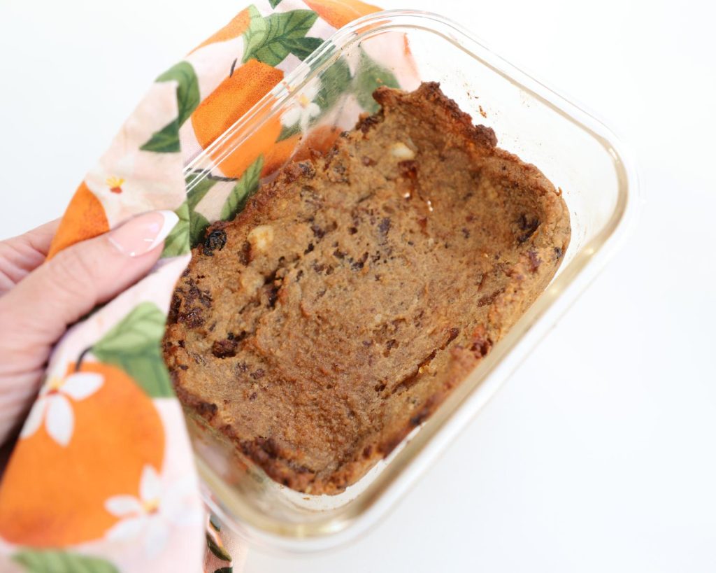 Overhead shot of a clear baking tray with a lightly brown hot cross bun pudding mix on the base, held by a yellow, orange, and green tea towel wrapped around a hand.
