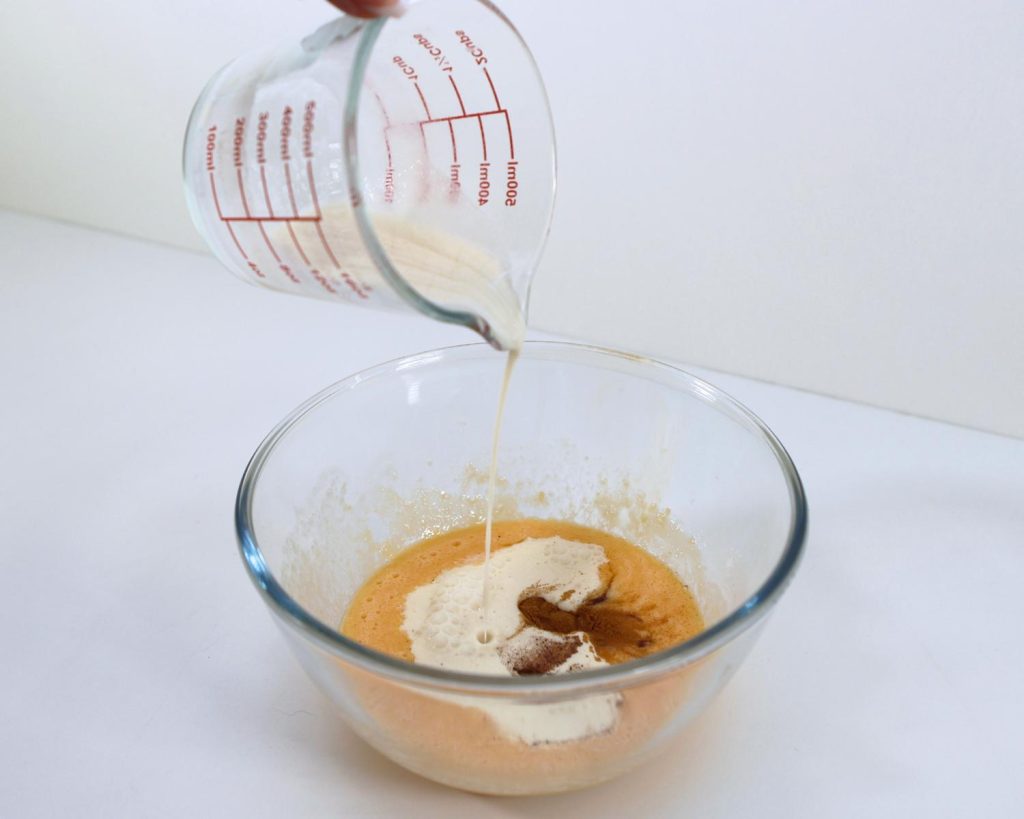 Clear glass mixing bowl on a white benchtop, half filled with hot cross bun custard pudding ingredients, as cream is poured in from a jug above.