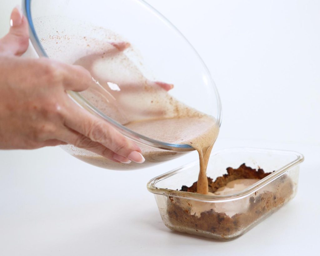 Clear glass baking tray on a white benchtop, half filled with a baked hot cross bun base, as custard mixture is poured in from a clear glass mixing bowl by two hands above. 