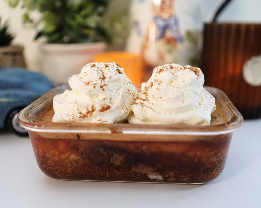 Clear glass baking tray on a white benchtop, filled with hot cross bun clear baking dish filled with hot cross bun custard pudding topped with whipped cream, a blurred multi-coloured background. 