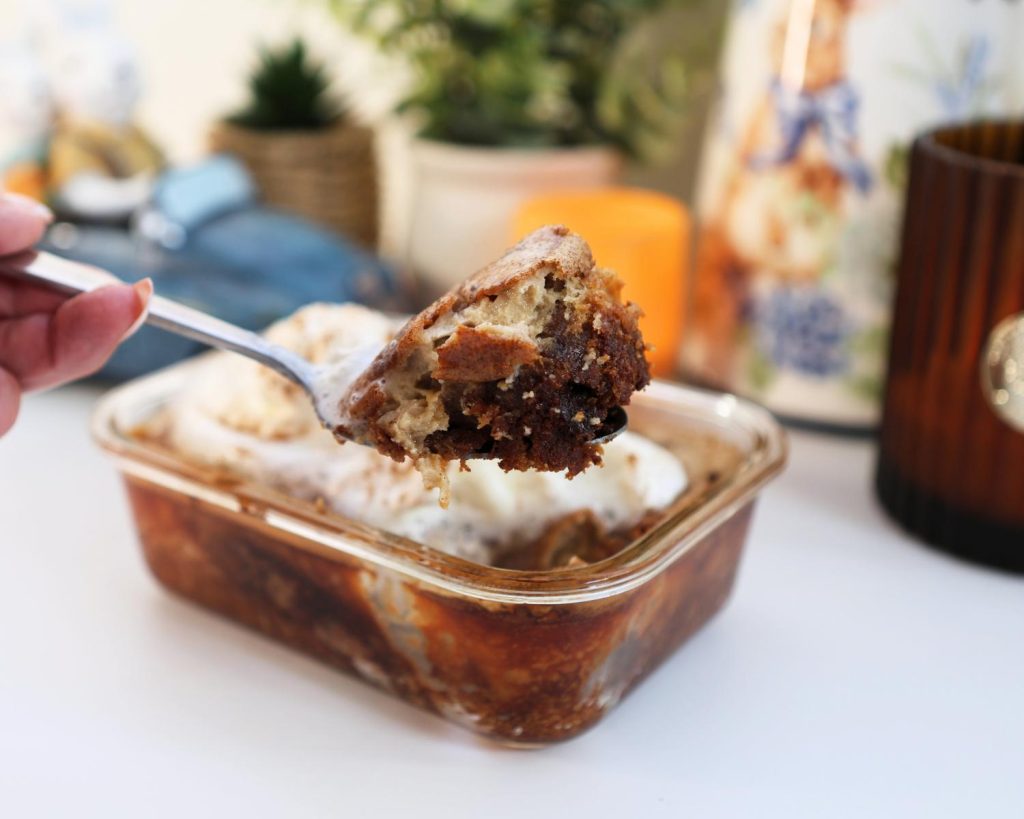 Shot of a hand holding a silver spoon with a scoop of hot cross bun custard budding in focus in front of a blurred background with a clear glass baking tray on a white benchtop, filled with hot cross bun clear baking dish filled with hot cross bun custard pudding topped with whipped cream. 