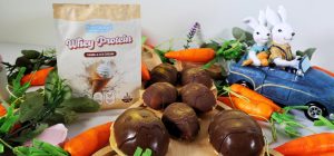 A yellow, blue, and white Whey Protein by Feel Good Protein (Vanilla Ice Cream) sample sachet beside 7 marshmallow easter eggs laid out across a wooden board, surrounded by an array of multicoloured easter decorations in the foreground and background.