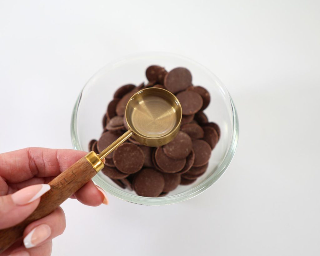Overhead view of a hand holding a wooden‑and‑metal tablespoon filled with clear liquid above a small glass bowl of chocolate melts on a white benchtop.