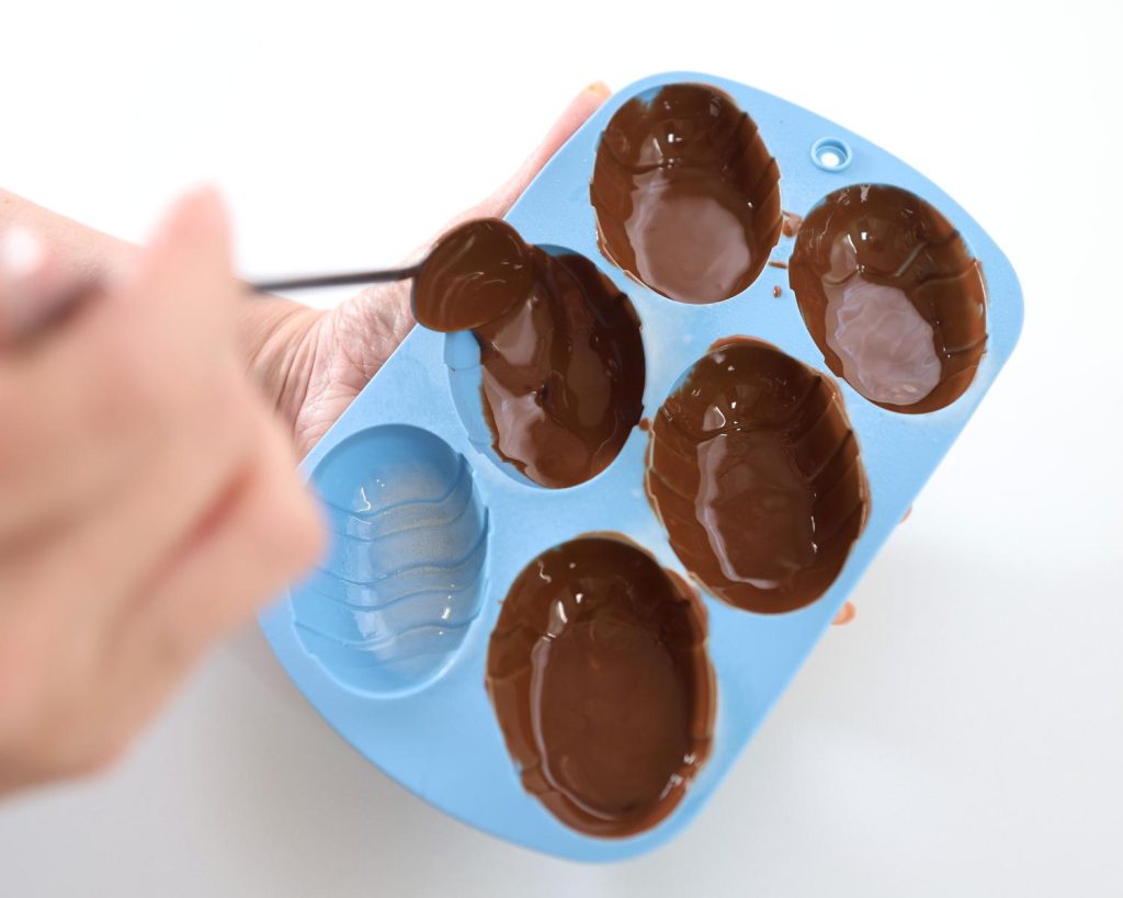 Overhead view of a blurred hand holding a metal and spoon drizzling chocolate into a bright blue silicone egg mould on a white benchtop.