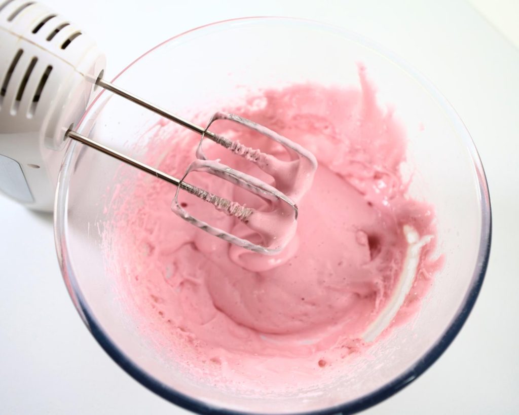 Overhead view of a white, two beater electric hand mixer next to a clear glass mixing bowl filled with bright pink marshmallow mixture.