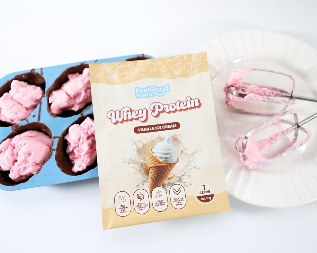 Overhead view of a yellow Whey Protein by Feel Good Protein (Vanilla Ice Cream) sachet resting on a blue egg mould tray filled with milk chocolate and bright pink marshmallows, next to a white plate holding two metal whisks coated in pink marshmallow.