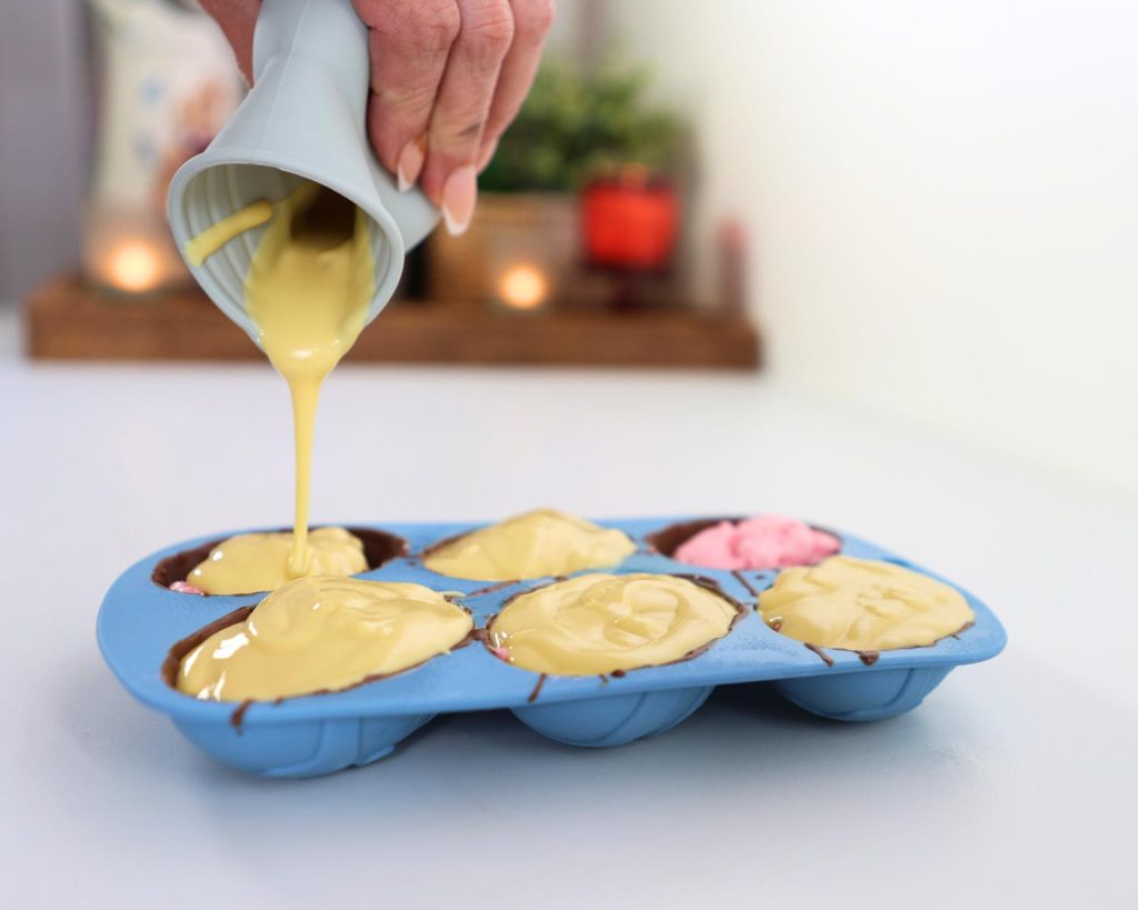 Close up photo of a hand pouring melted white chocolate from a blue jug into a bright blue silicone egg mould tray on a white benchtop.