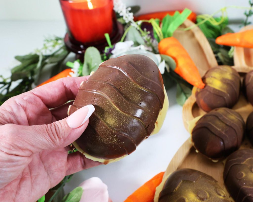 Close up photo of a hand holding a Marshmallow Easter Egg front of a blurred background of Easter decorations and a tray of Marshmallow Easter Eggs.
