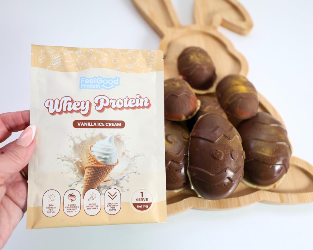 Point of view shot of a hand holding a yellow sample sachet of Whey Protein by Feel Good Protein (Vanilla Ice Cream) in front of a rabbit-shaped wooden board of Marshmallow Easter Eggs.