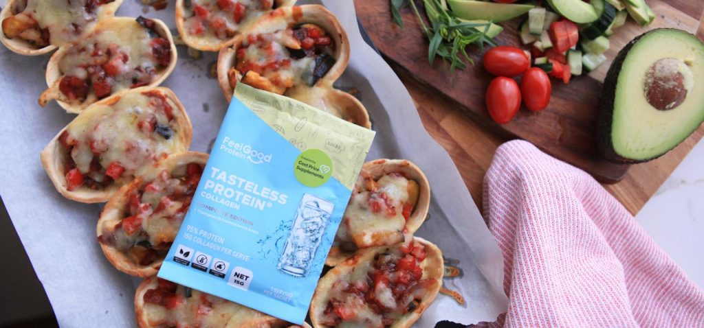 Overhead shot of a blue and yellow Tasteless Protein Collagen by Feel Good Protein sample sachet on top of a baking tray lined with two rows of Salsa Chicken Taco Boats, next to two wooden boards with an assortment of bright vegetables on top.