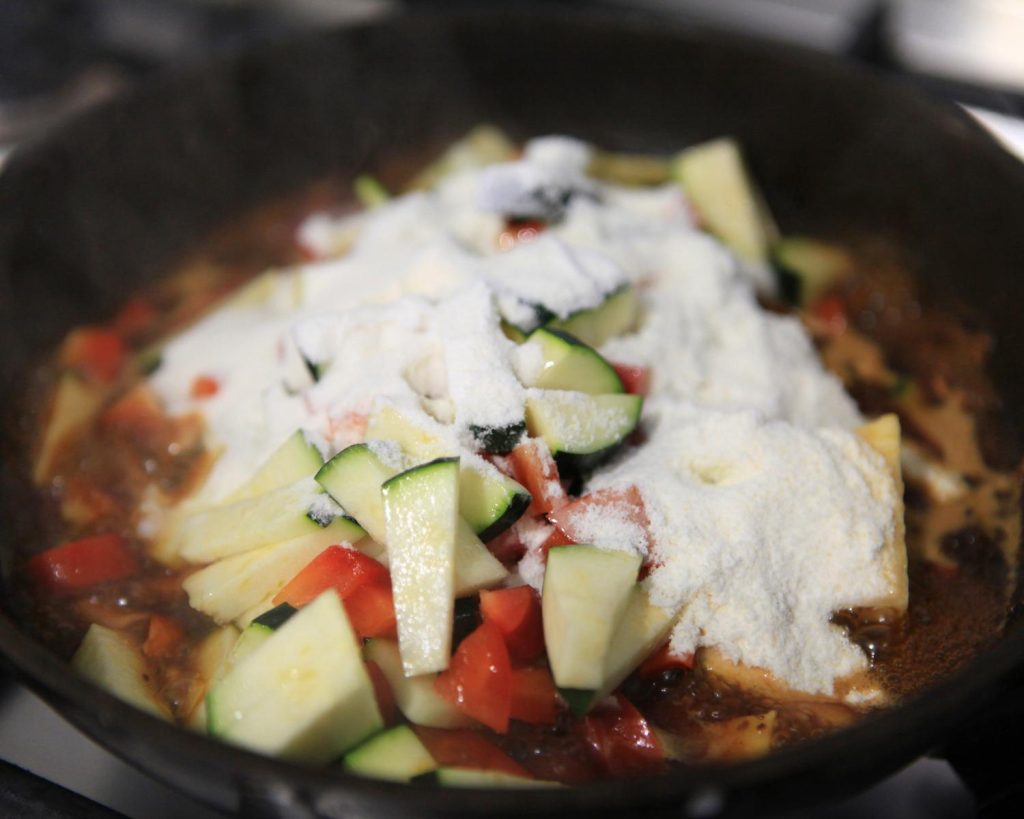 Point of view shot of a black cooking pan filled halfway with bright green and red chopped vegetables with a pile of white protein powder covering the pile. 