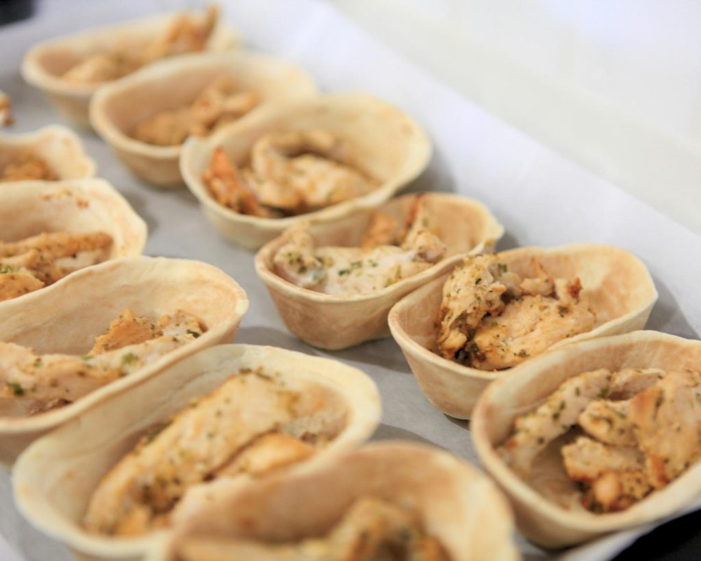 Overhead shot of 12 taco boats lined in two rows on a baking tray, half filled with cooked seasoned chicken strips. 