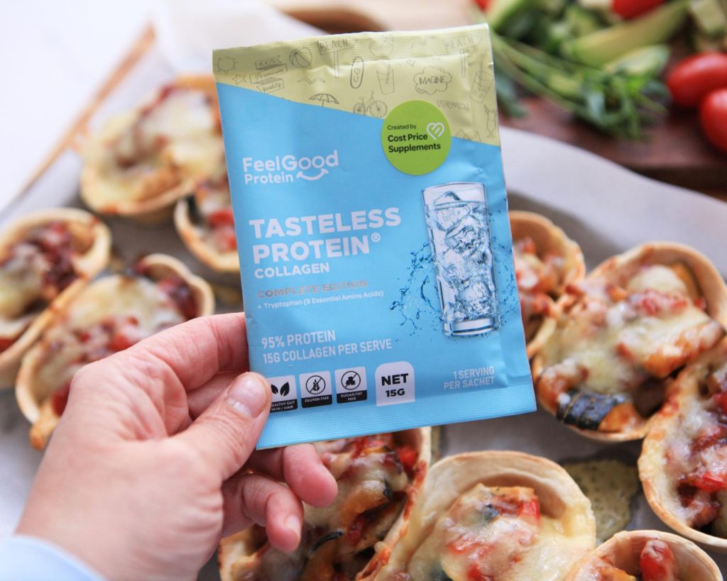 Point of view shot of a hand holding a blue and yellow Tasteless Protein Collagen by Feel Good Protein sample sachet in front of a blurred background featuring a baking tray lined with two rows of Salsa Chicken Taco Boats covered in crispy melted cheese, next to two wooden boards with an assortment of bright vegetables. 