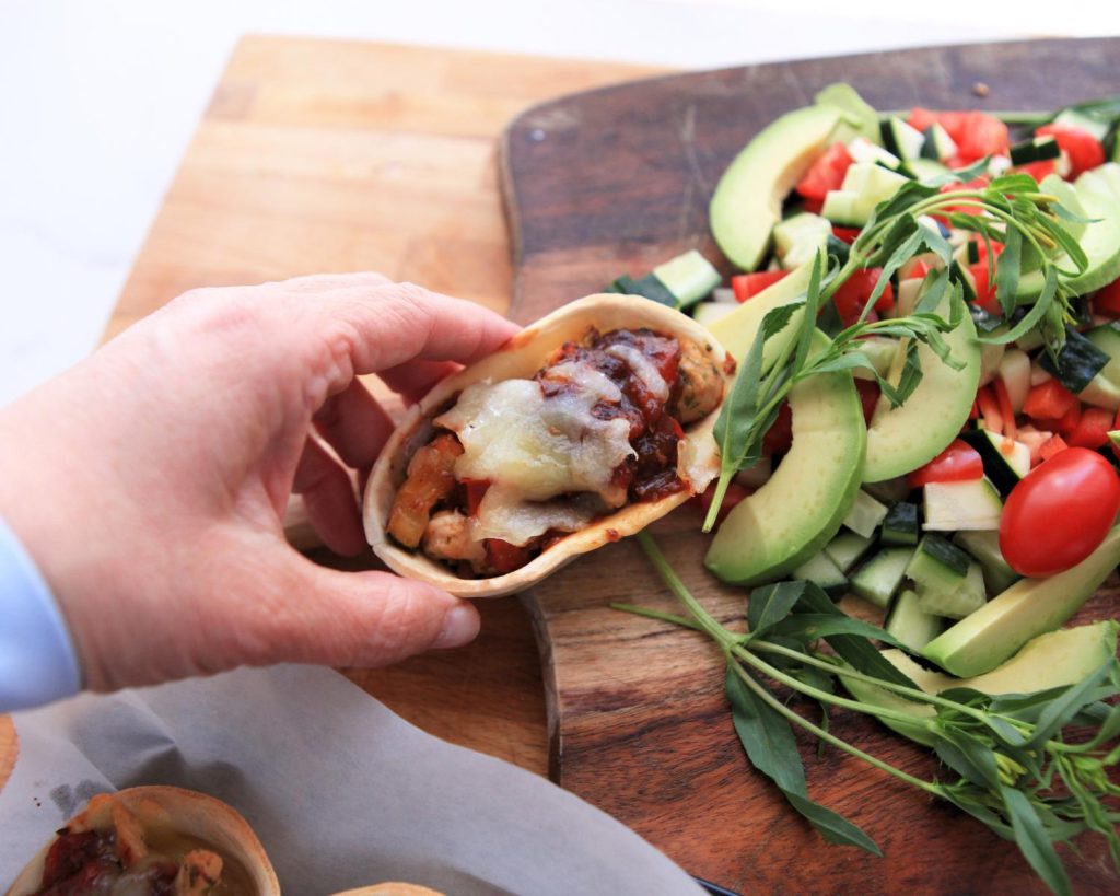 Point of view shot of a hand holding one Salsa Chicken Taco Boats covered in crispy melted cheese over a background with two wooden boards and an assortment of bright vegetables. 