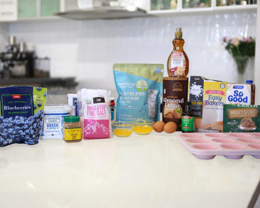 Front-facing photo showing all the ingredients for blueberry muffins with crumble topping arranged neatly on a marble benchtop, with a softly blurred modern kitchen in the background.