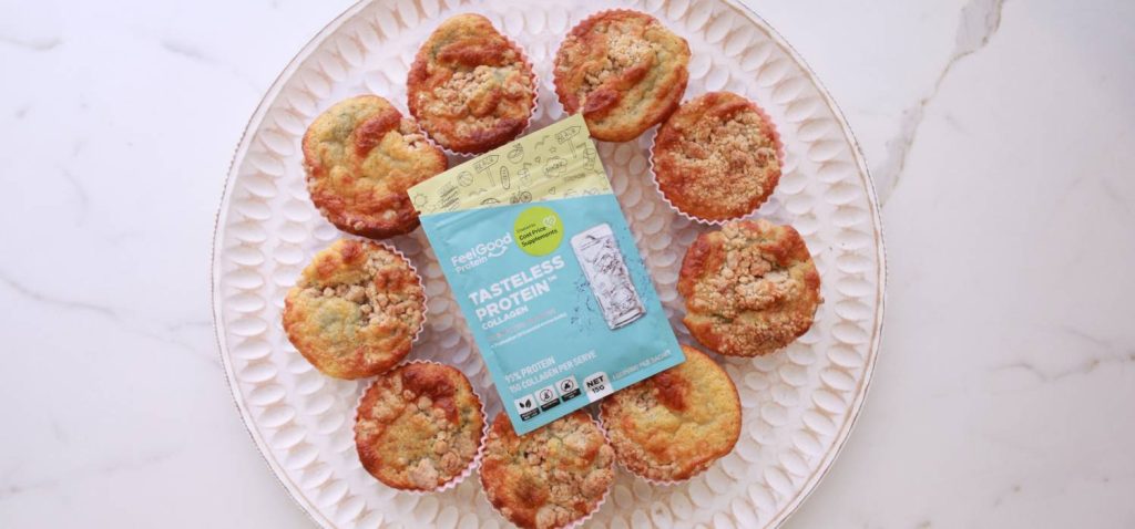 Top‑down view of a Feel Good Protein Tasteless Protein Collagen sachet in blue and yellow packaging centred on a white plate with eight muffins arranged around it.