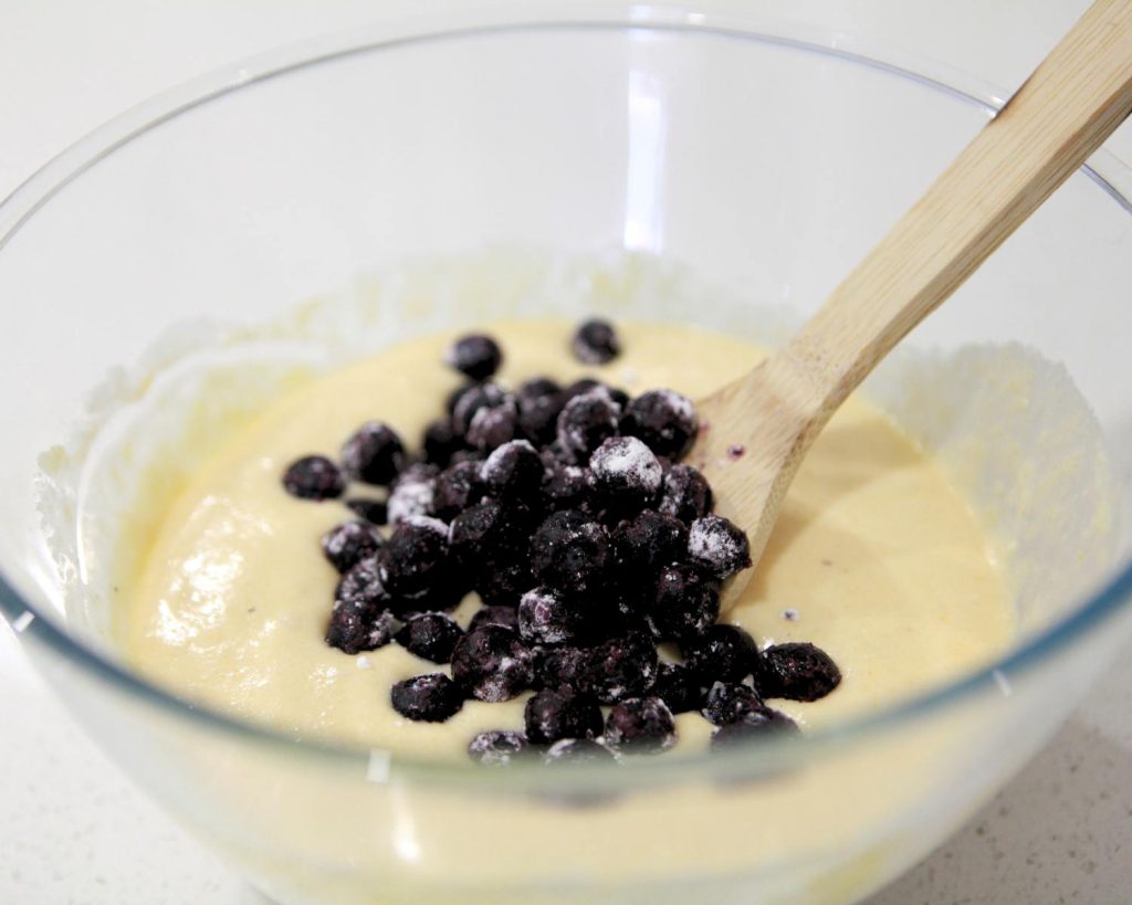 Photo of a clear glass mixing bowl half‑filled with muffin batter with blueberries in the centre, with a wooden spoon resting upright on the rim. 
