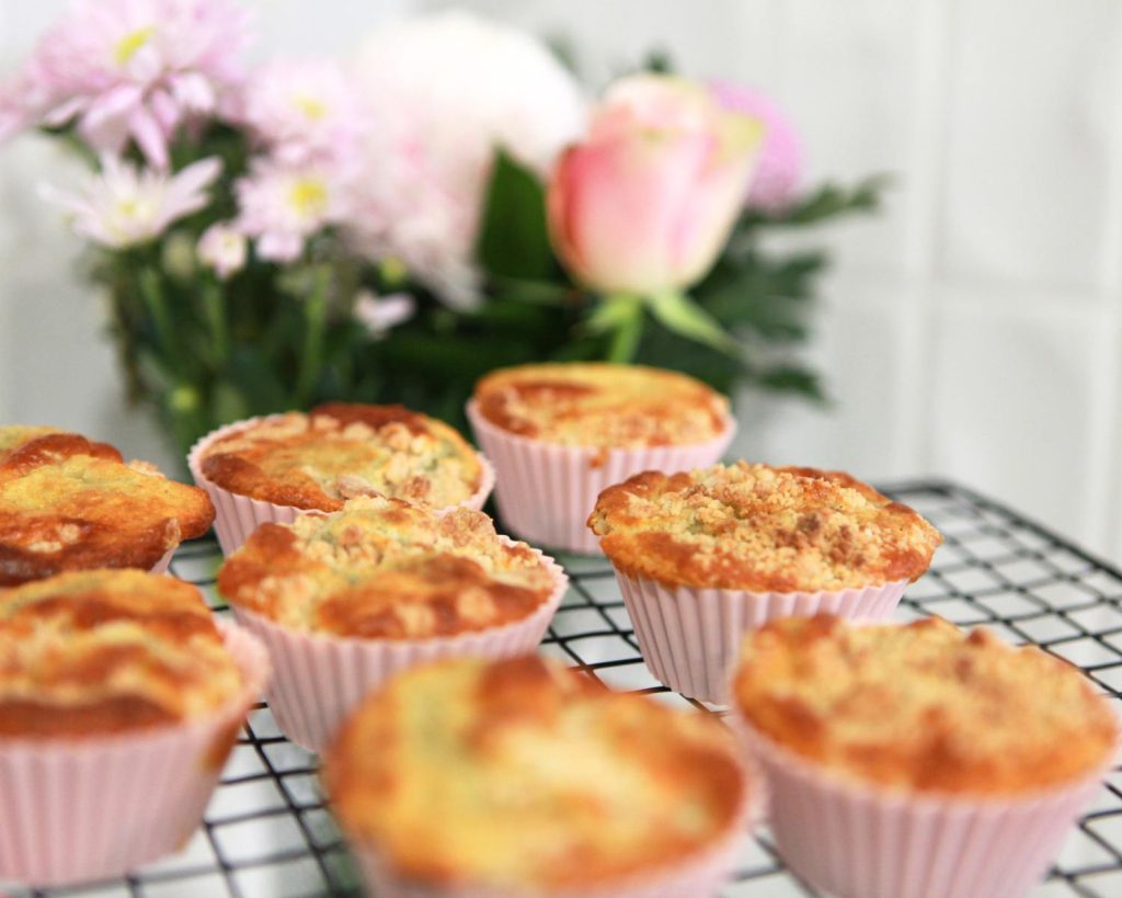 Golden yellow muffins in pink paper cases cooling on a black wire rack with soft pink flowers blurred in the background.
