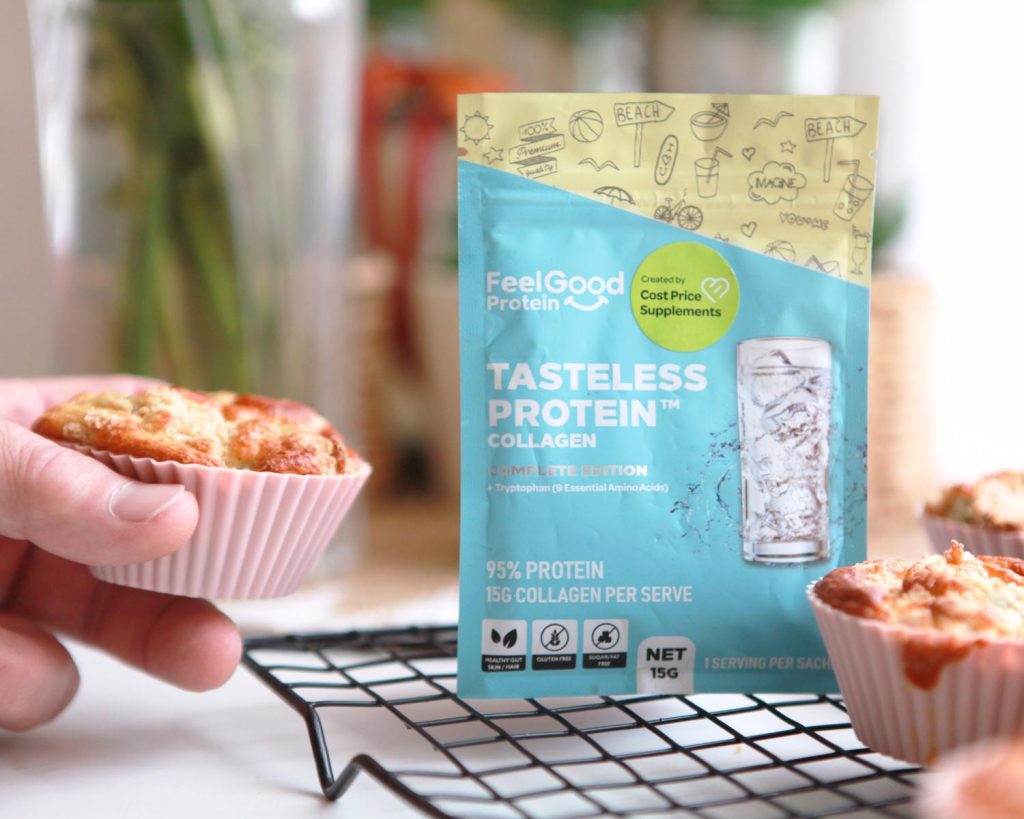 Top‑down view of a Feel Good Protein Tasteless Protein Collagen sachet in blue and yellow packaging on a black wire cooling rack beside freshly baked muffins, with a hand lifting one muffin.