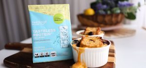 Front on photo of a yellow, blue, and white Tasteless Protein Collagan by Feel Good Protein sample sachet beside 2 ramekins filled with Blueberry French Toast, laid out across a wooden board in front of a blurred kitchen benchtop background.