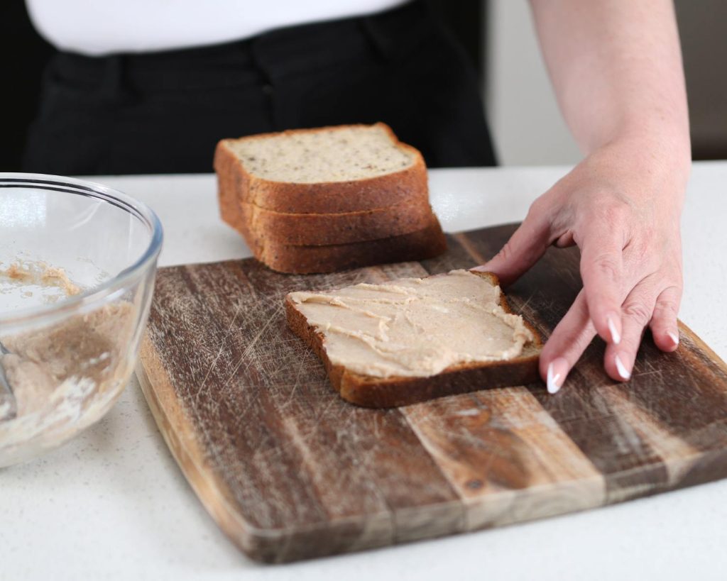 Photo of a hand spreading a cream cheese mixture onto a slice of bread on a wooden board, which sits on a kitchen benchtop beside a clear mixing bowl and in front of a stack of bread.