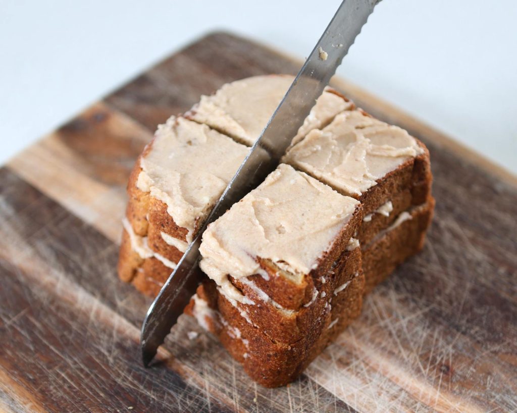 Photo of a knife cutting 4 pieces of stacked bread with cream cheese spread on a wooden board, which sits on a kitchen benchtop. 