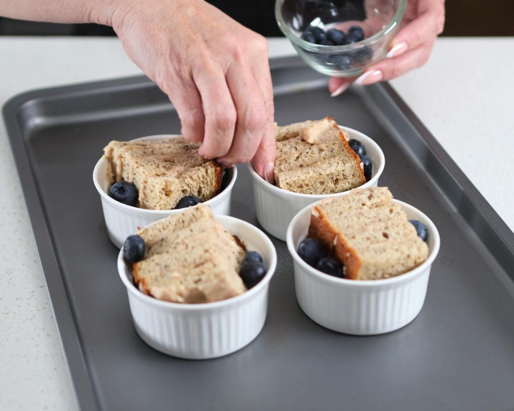Photo of a hand holding a small bowl of blueberries, sprinkling them into 4 ramekins on a baking tray.