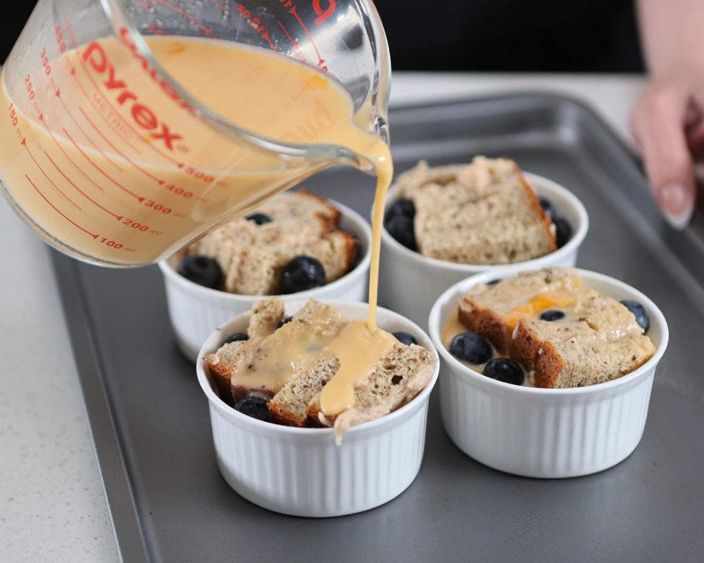 Photo of a clear jug of yellow liquid being poured into 4 ramekins on a baking tray.