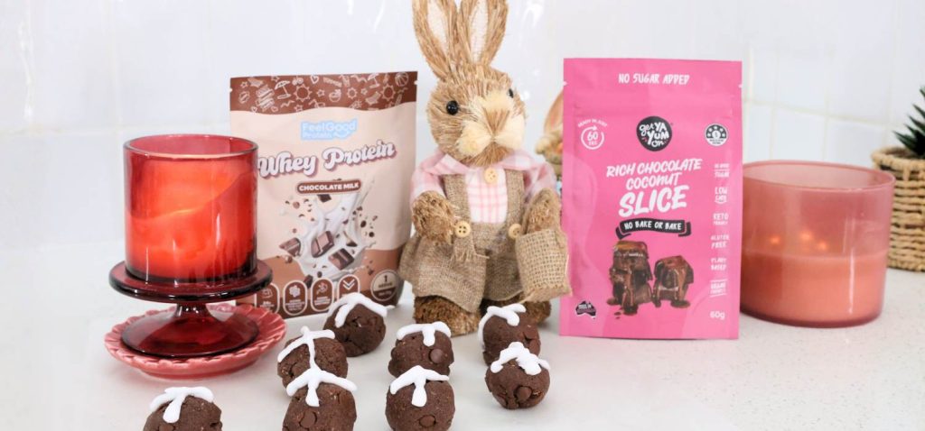 Front on photo of Whey Protein by Feel Good Protein (Chocolate Milk) sachet and a Rich Chocolate Coconut Slice Mix by Get Ya Yum On sachet on both sides of a Easter bunny statue centred behind 8 protein balls.